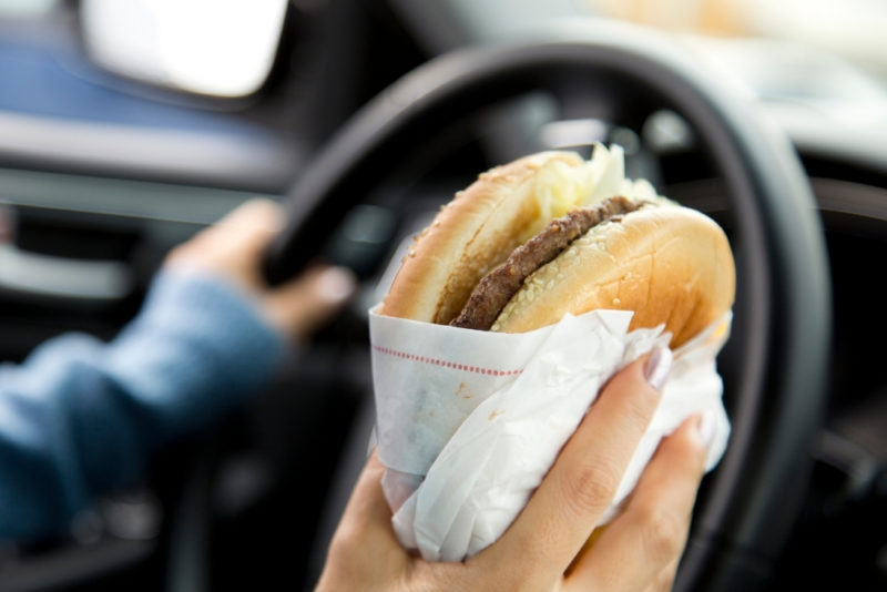 It's hard to get true nourishment from your food when you are eating on the run. Photo of someone holding a burger in one hand while driving with the other.