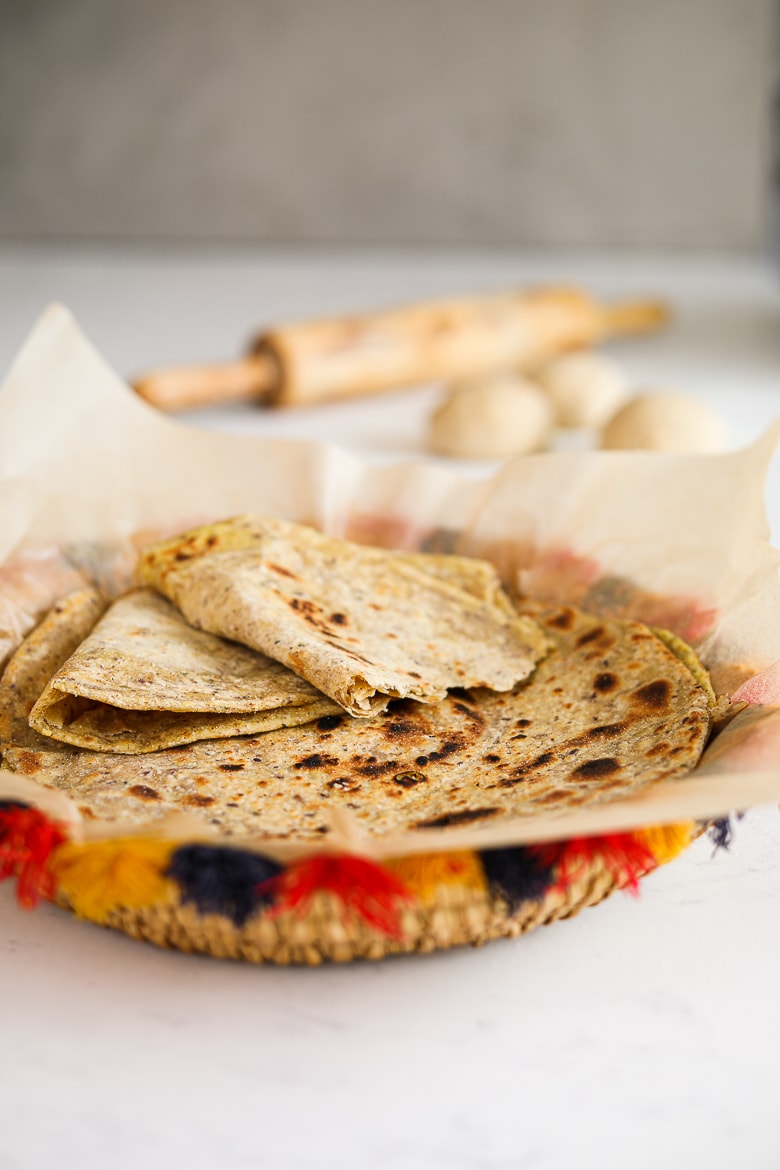 a colourful traditional South Asian straw basket containing a pile of parathas (Indian flat bread) with one paratha ripped in half. A rolling pin and balls of dough in the background
