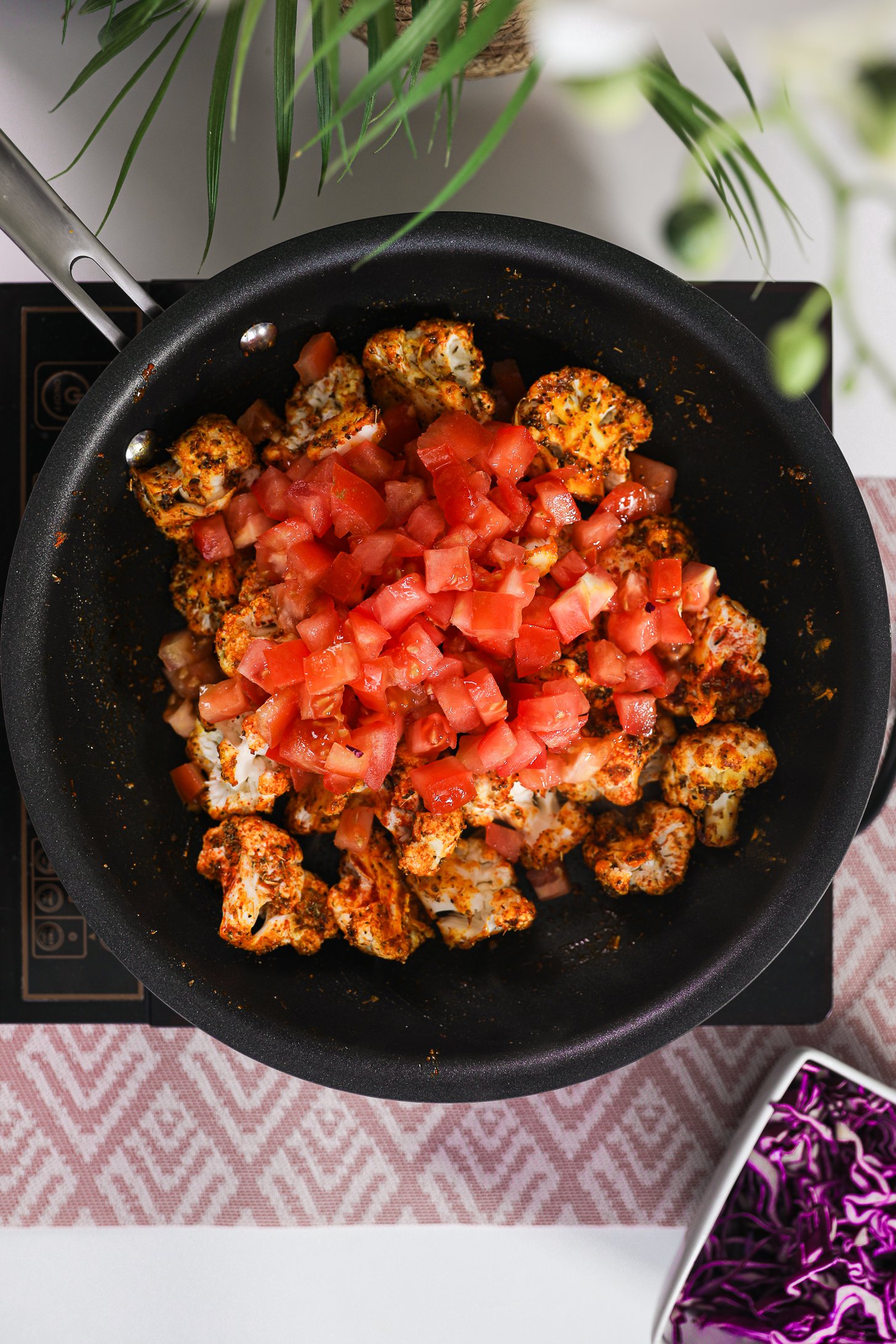 Pan of spice-coated cauliflower florets topped with chopped tomatoes on a mobile worktop.